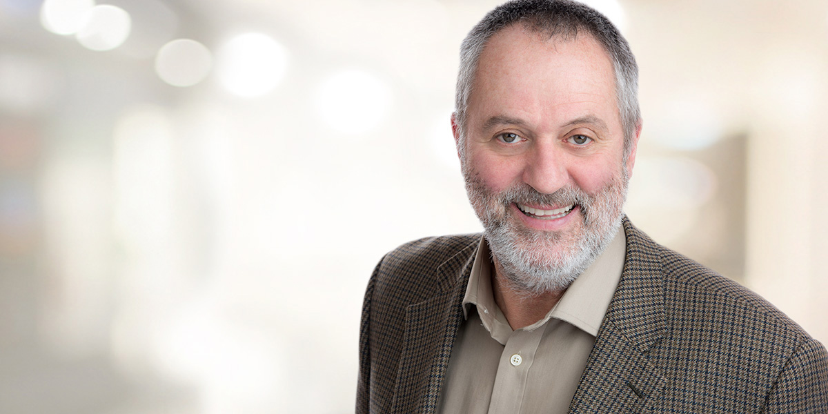 Beareded gentleman in a tweed jacketa standing gainst a company colour branded backdrop, smiles for his Headshots made at Yungblut's Downtown St. Catharines Studio 